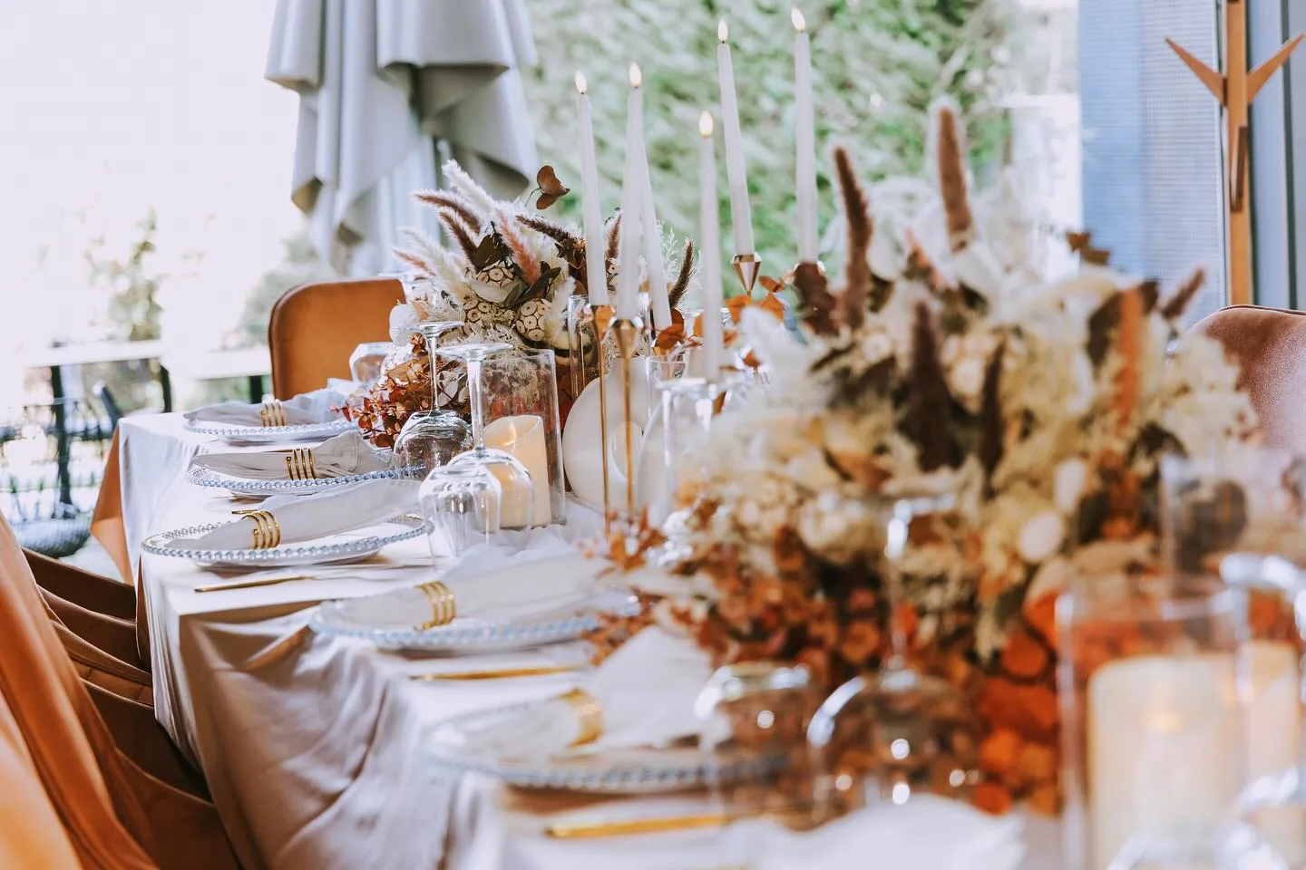 Long banquet table set with beaded charger plates, gold cutlery, tall candles and dried-flower arrangements