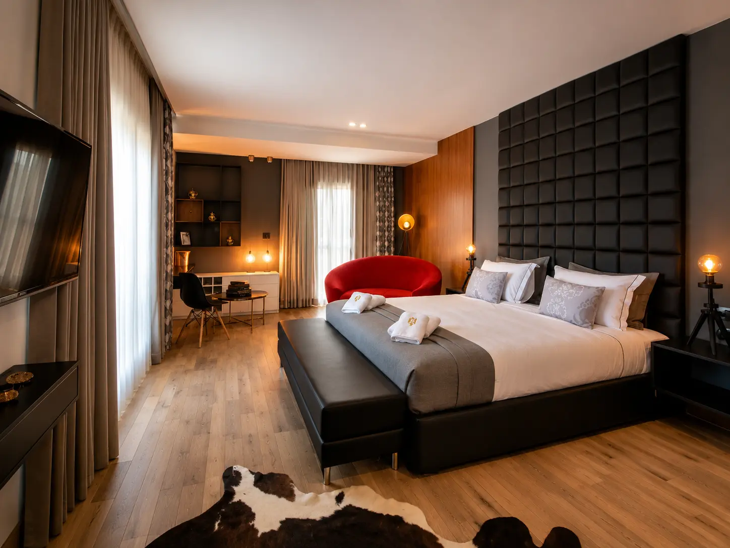Guest room with a king bed, tall padded grey headboard, red curved sofa and a cowhide rug on wood flooring