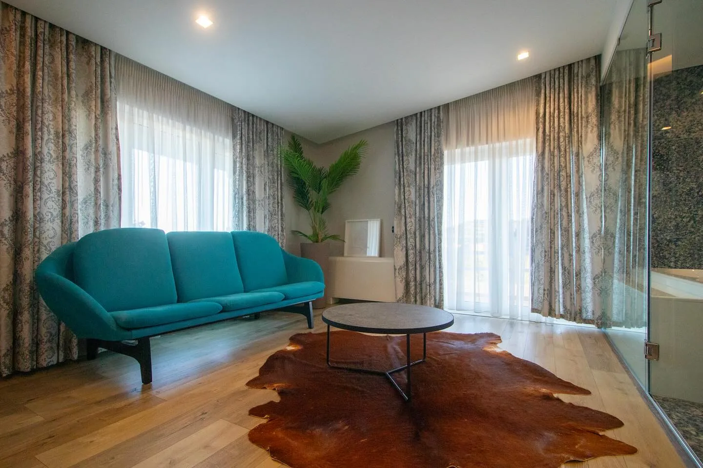 Suite sitting area with a teal three-seat sofa, round stone table and potted palm beside a glass wall